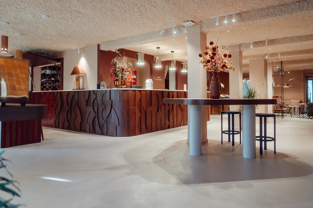 Salle d’accueil de la Maison Melba avec bar, piliers et tables hautes, sol en béton ciré